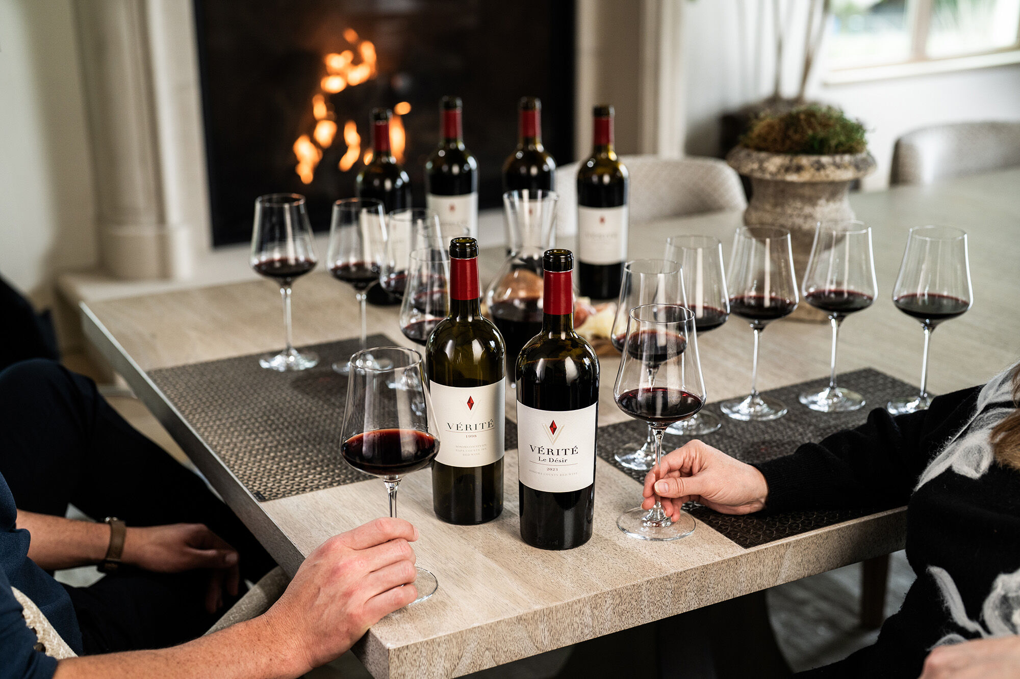 9 wine glasses on a light marble table with wine bottles, a fireplace in the background, and two people's hands holding wine glasses