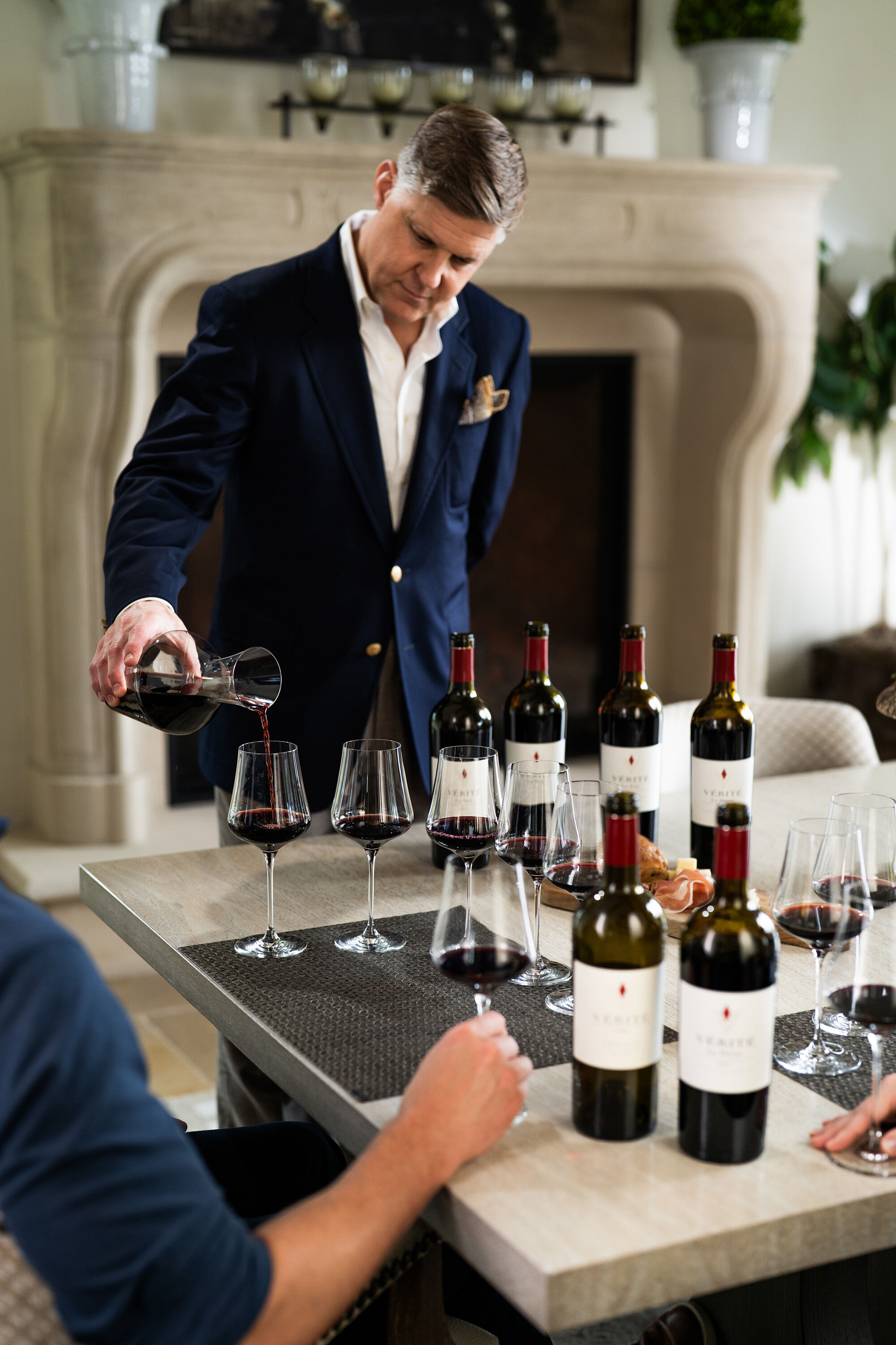 A man pouring red wine into a wine glass, with seven other wine glasses and six bottles surrounding on the table.