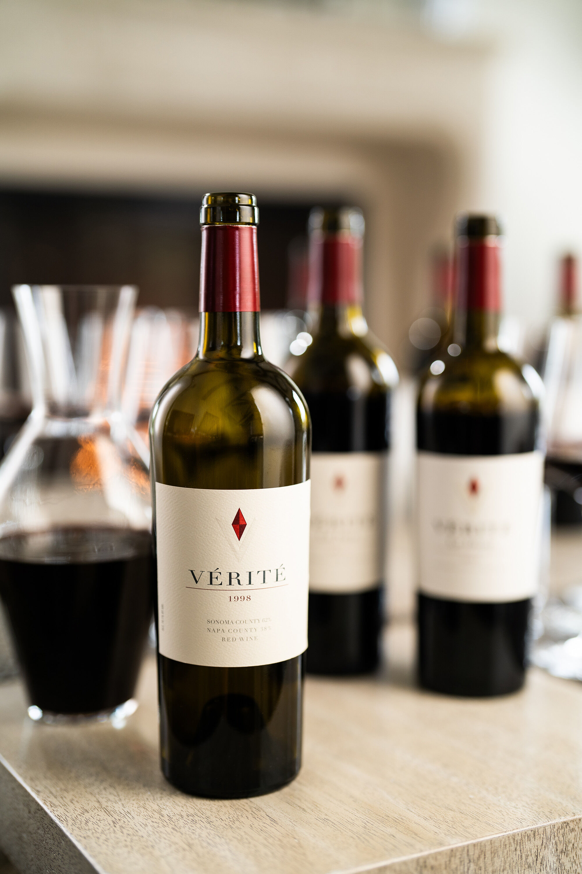 A close up of a bottle of Verite wine with the 1998 vintage on the label, and two wine bottles and a decanter blurred in the background.