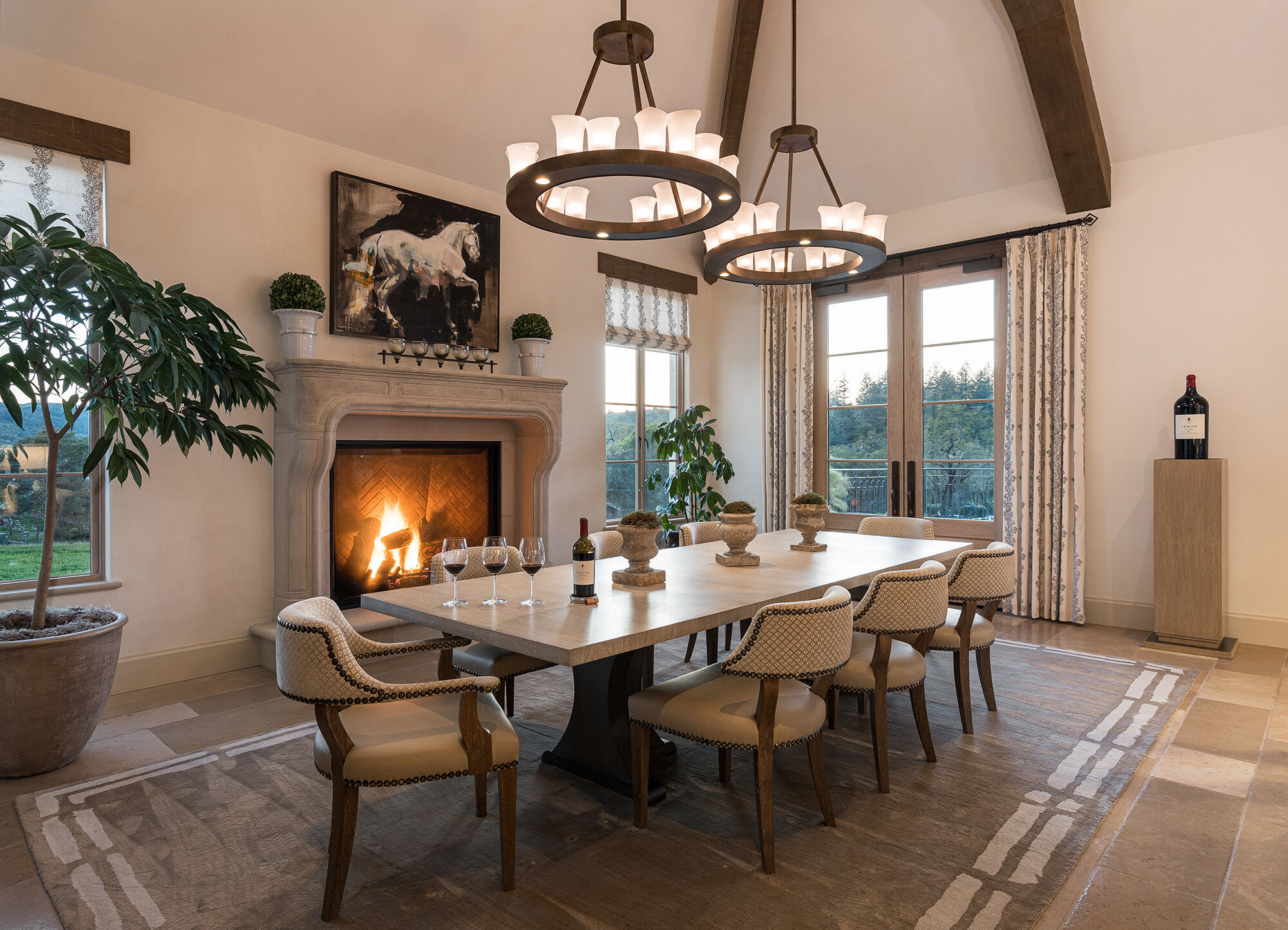 An 8-person table with light beige chairs in front of a fireplace, with a wine bottle and three glasses.