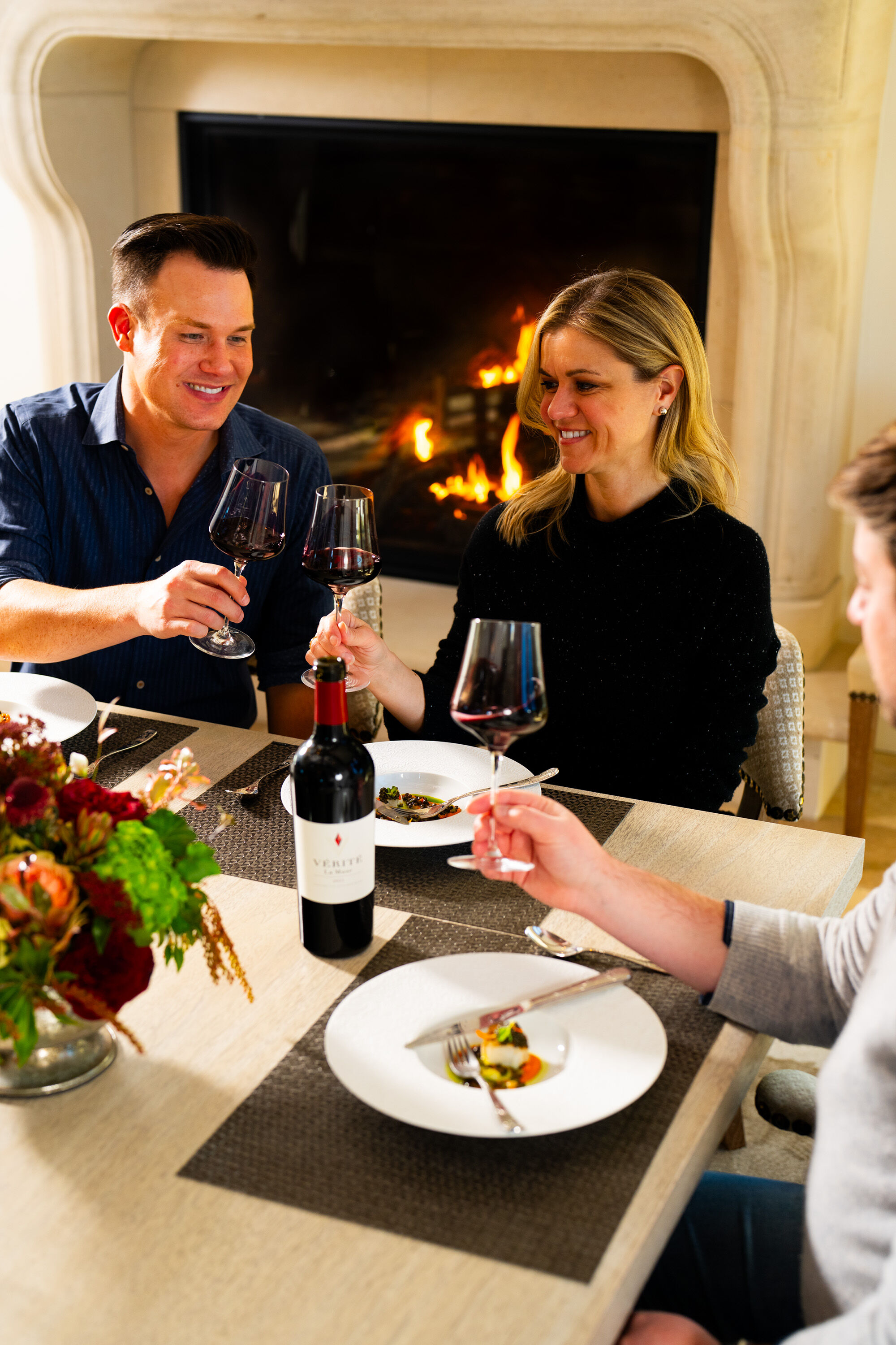 Three people around a dining table in front of a fireplace, holding wine glasses, smiling and clinking glasses.